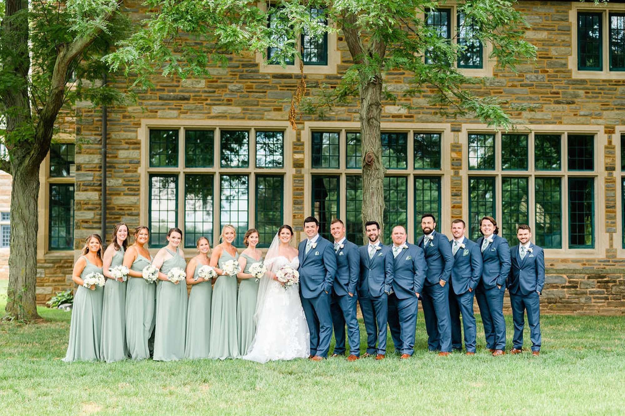 newlyweds stand with bridesmaids in sage green gowns and groomsmen in navy suits