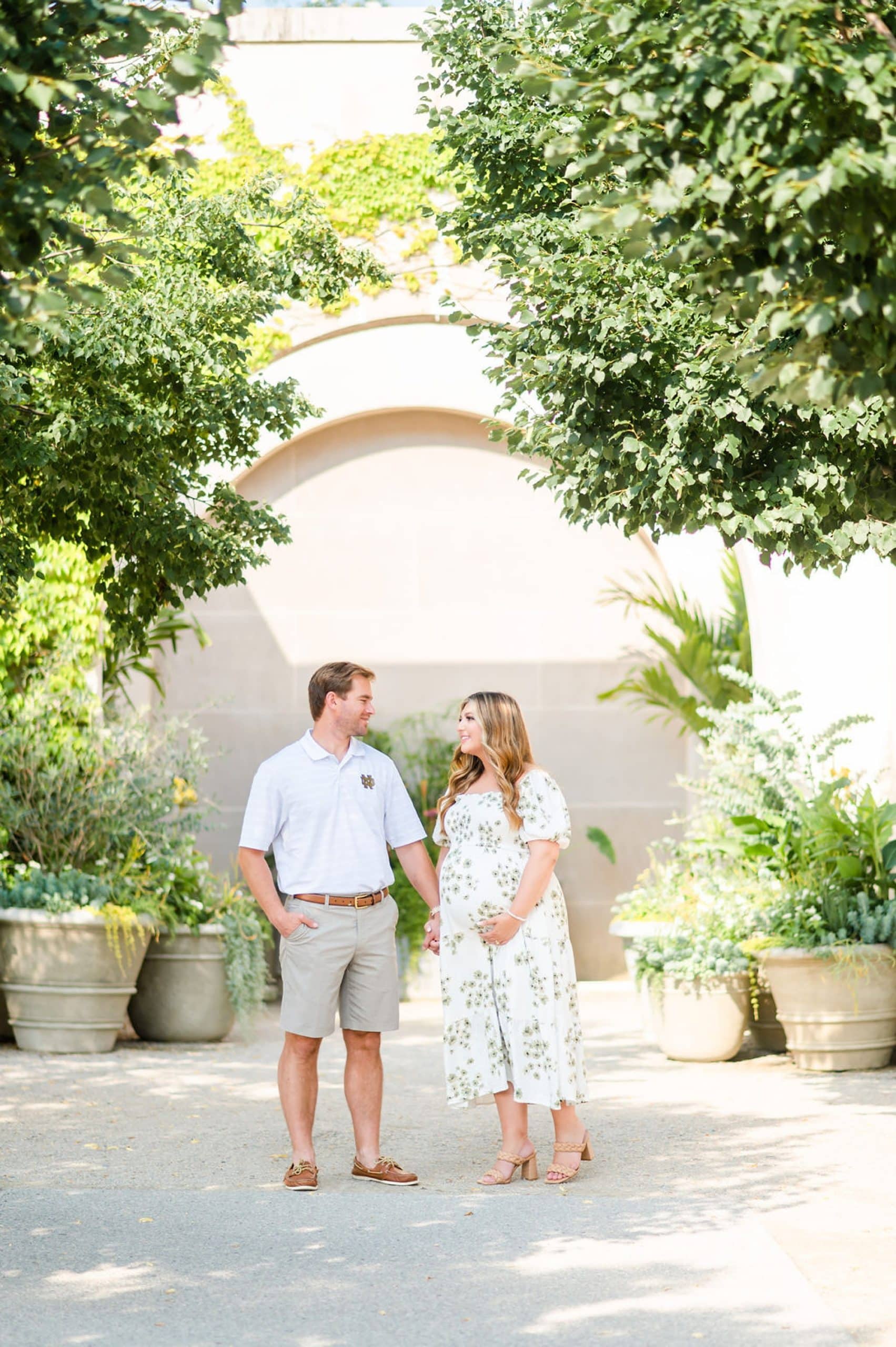 husband and wife pose by tan wall during Longwood Gardens maternity portraits
