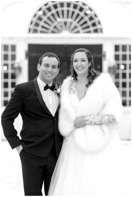 black and white photo of Bride and groom stand infront of a black arched door in a snow storm