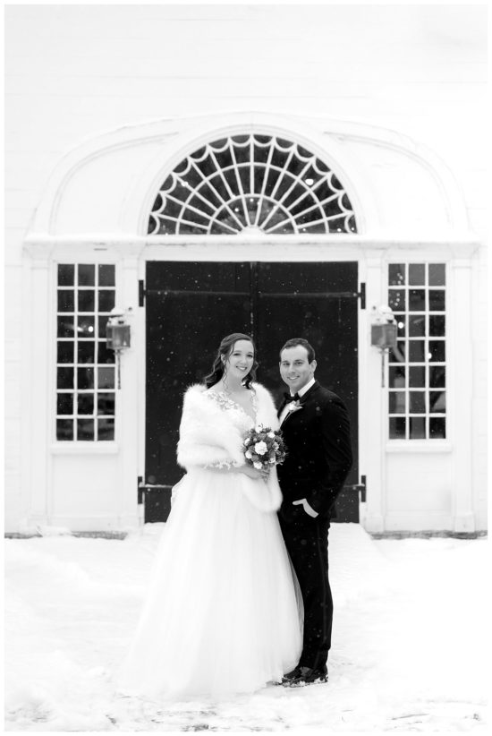 Bride and groom stand infront of a black arched door in a snow storm